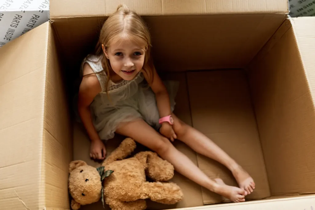 Kleines Mädchen sitzt mit Teddybär in einem Umzugskarton und spielt fröhlich.